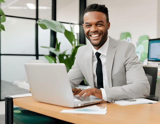 A businessman sits at a desk, typing on his laptop, with a broad smile, wearing a crisp suit, surrounded by office decor and natural light.